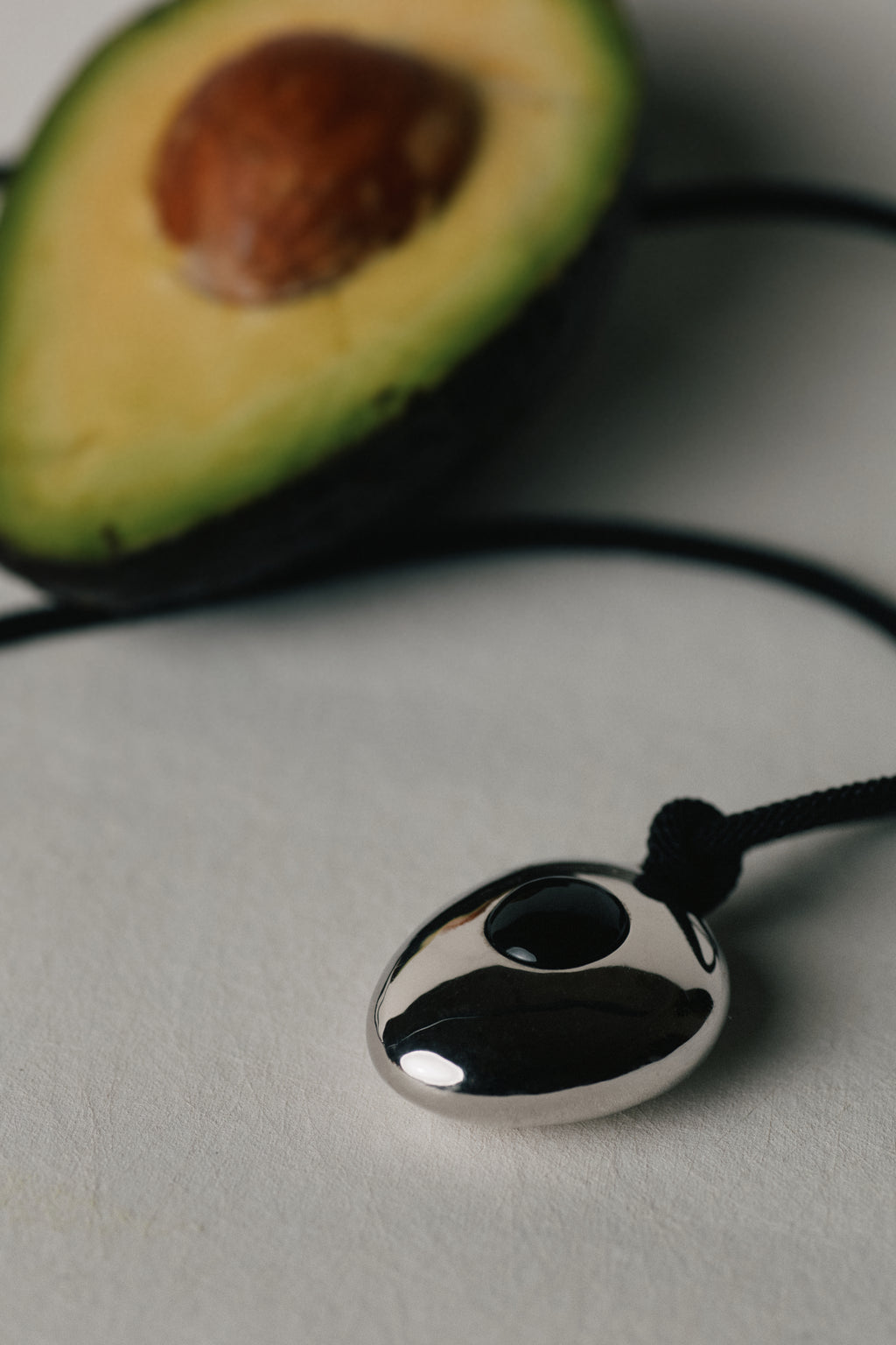 Avocado Mirror Pendant Necklace — Silver pendant on a cord with an avocado cut in half in the background