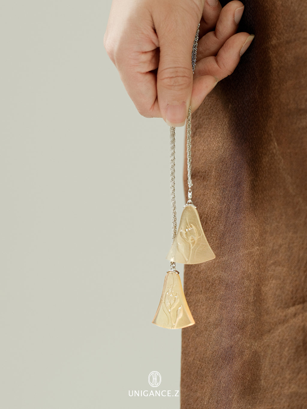 Gold bell-shaped pendant on a chain held by a hand against a neutral background