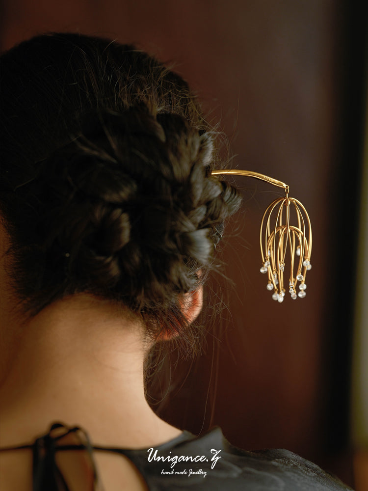 Drooping Begonia Hairpin — Close-up of a gold earring worn by a person with braided hair against a dark background.