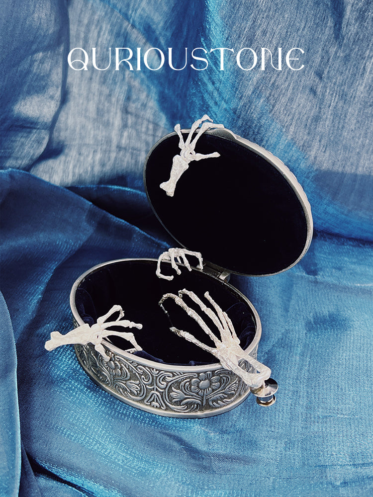 Gold Digger · Long Claw Earring (Single) — Silver skeleton hand earrings in an open jewelry box on a blue fabric background
