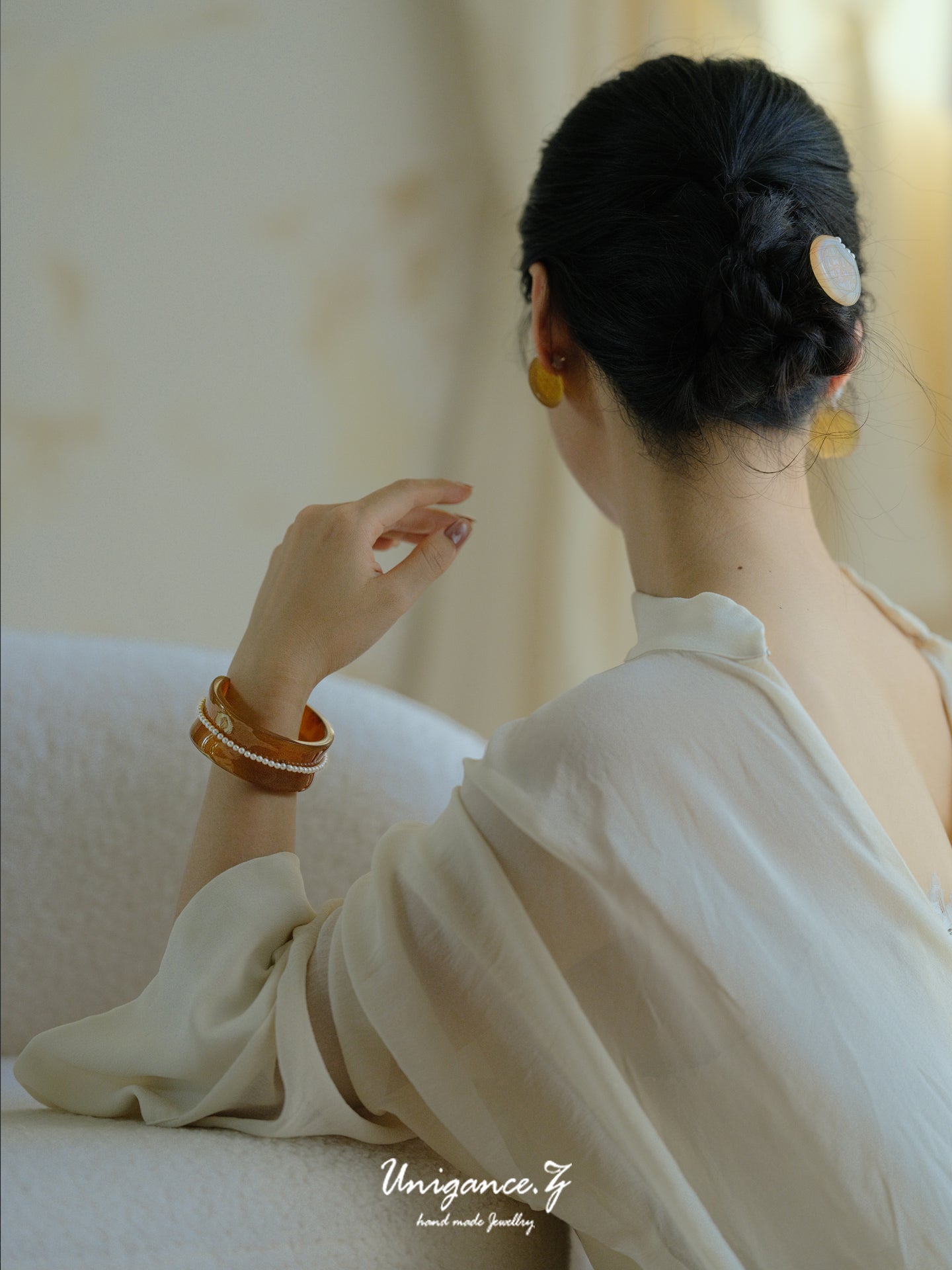 Woman in a white saree with gold jewelry sitting on a white couch.
