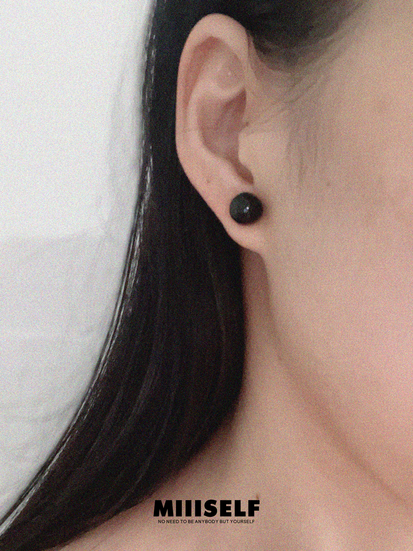 Close-up of an ear wearing a black stud earring with 'MIIISELF' branding.