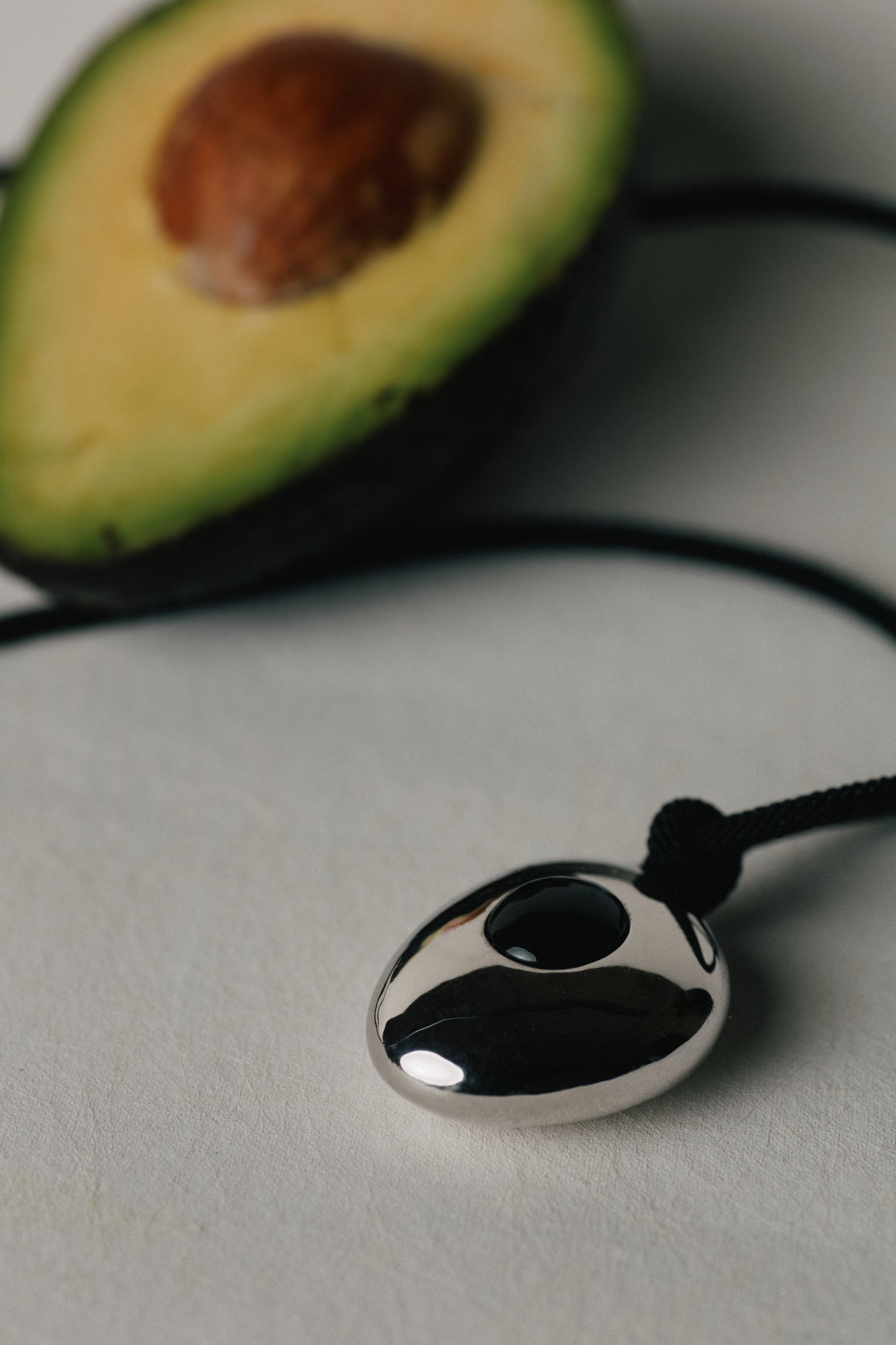 Silver pendant on a cord with an avocado cut in half in the background