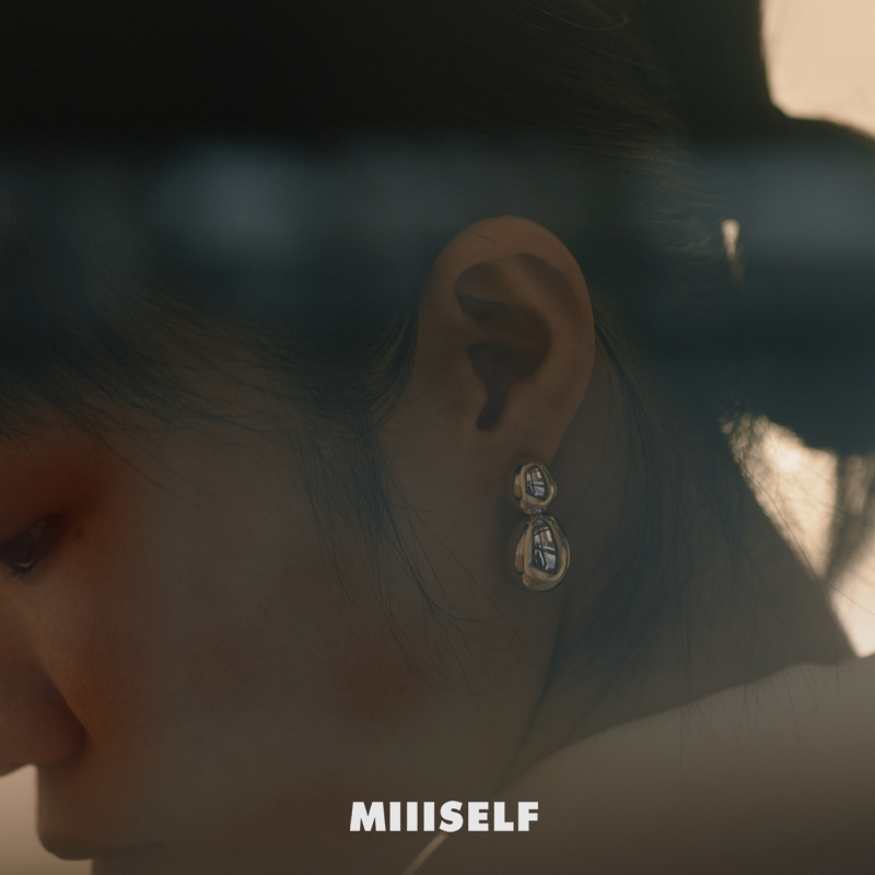Close-up of a person wearing gold earrings with 'MIIISELF' branding.
