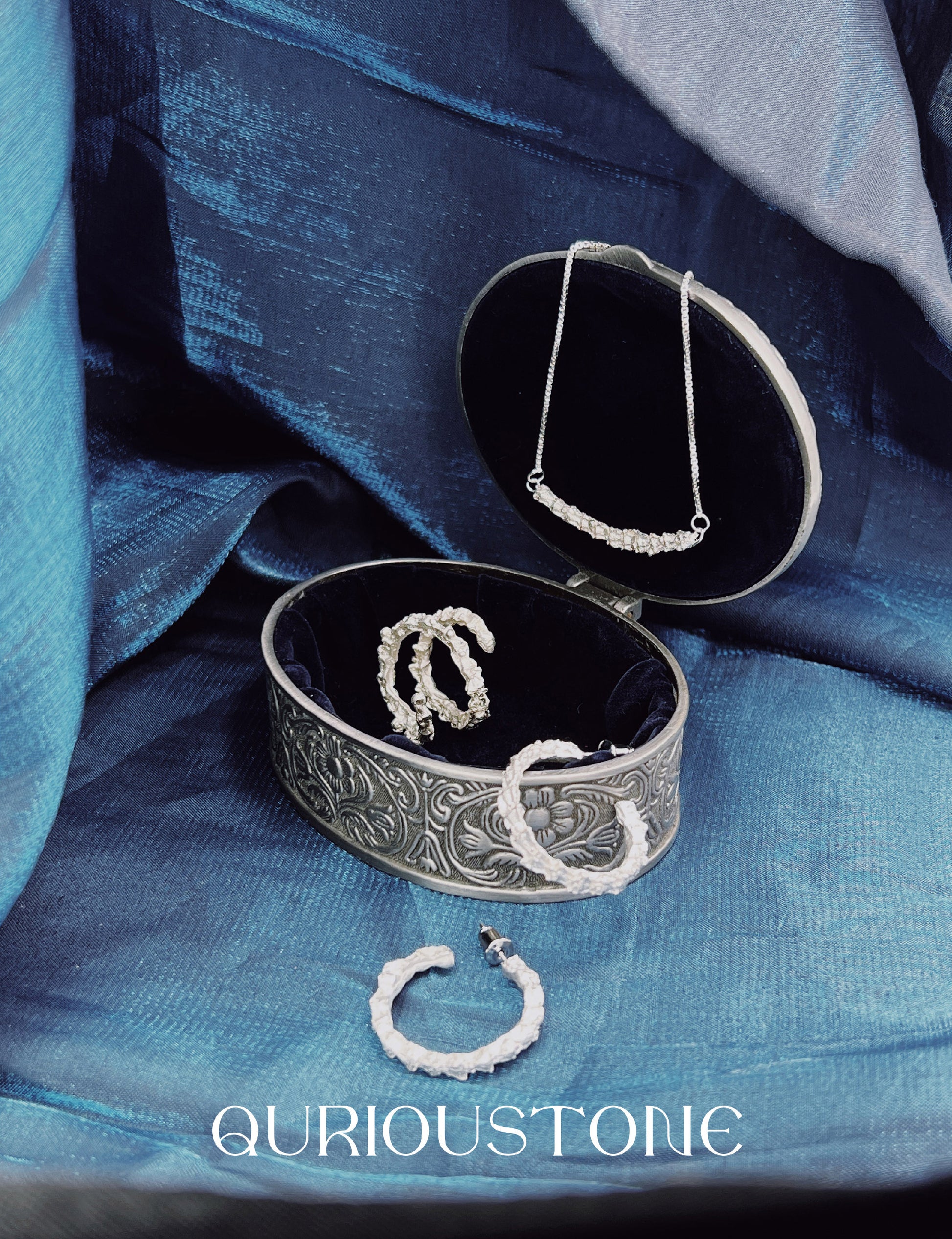 Jewelry box with necklaces on a blue fabric background, featuring the brand 'Quirous Stone'.