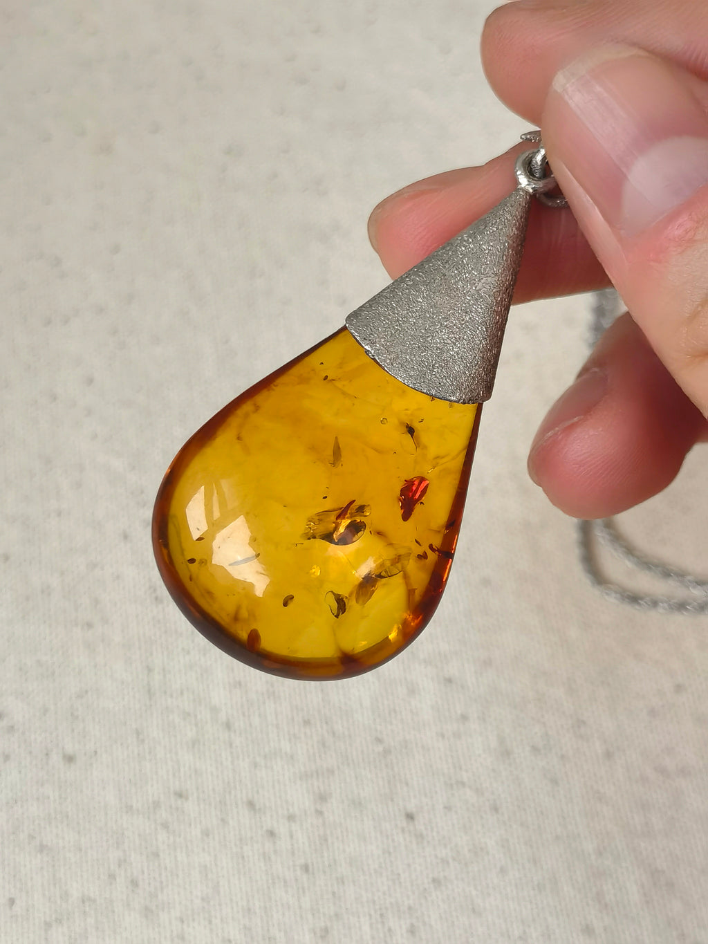 Natural Floral Amber & Matte Silver Necklace — Teardrop-shaped amber pendant with silver accent held by a hand on a neutral