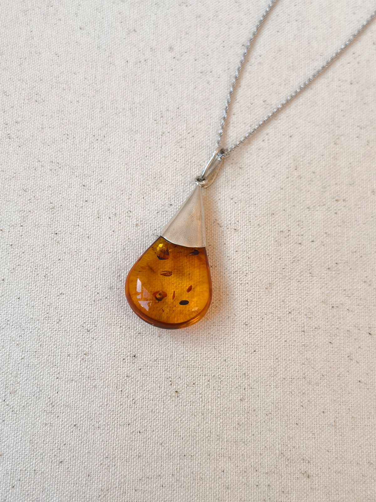 Natural Floral Amber & Matte Silver Necklace — Teardrop-shaped amber pendant on a chain against a beige fabric background