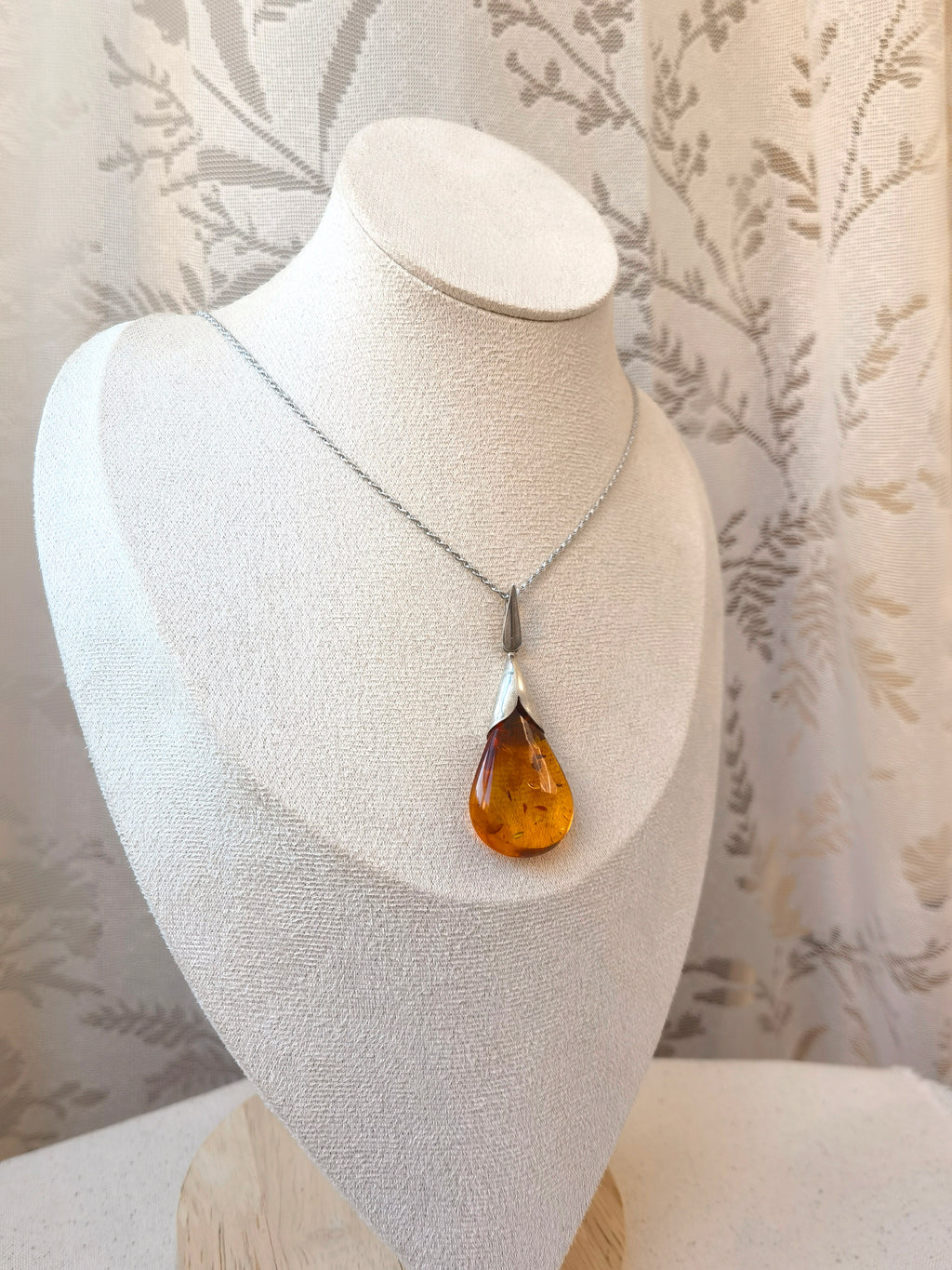 Natural Floral Amber & Matte Silver Necklace — Necklace with a teardrop-shaped amber pendant on a white bust against a