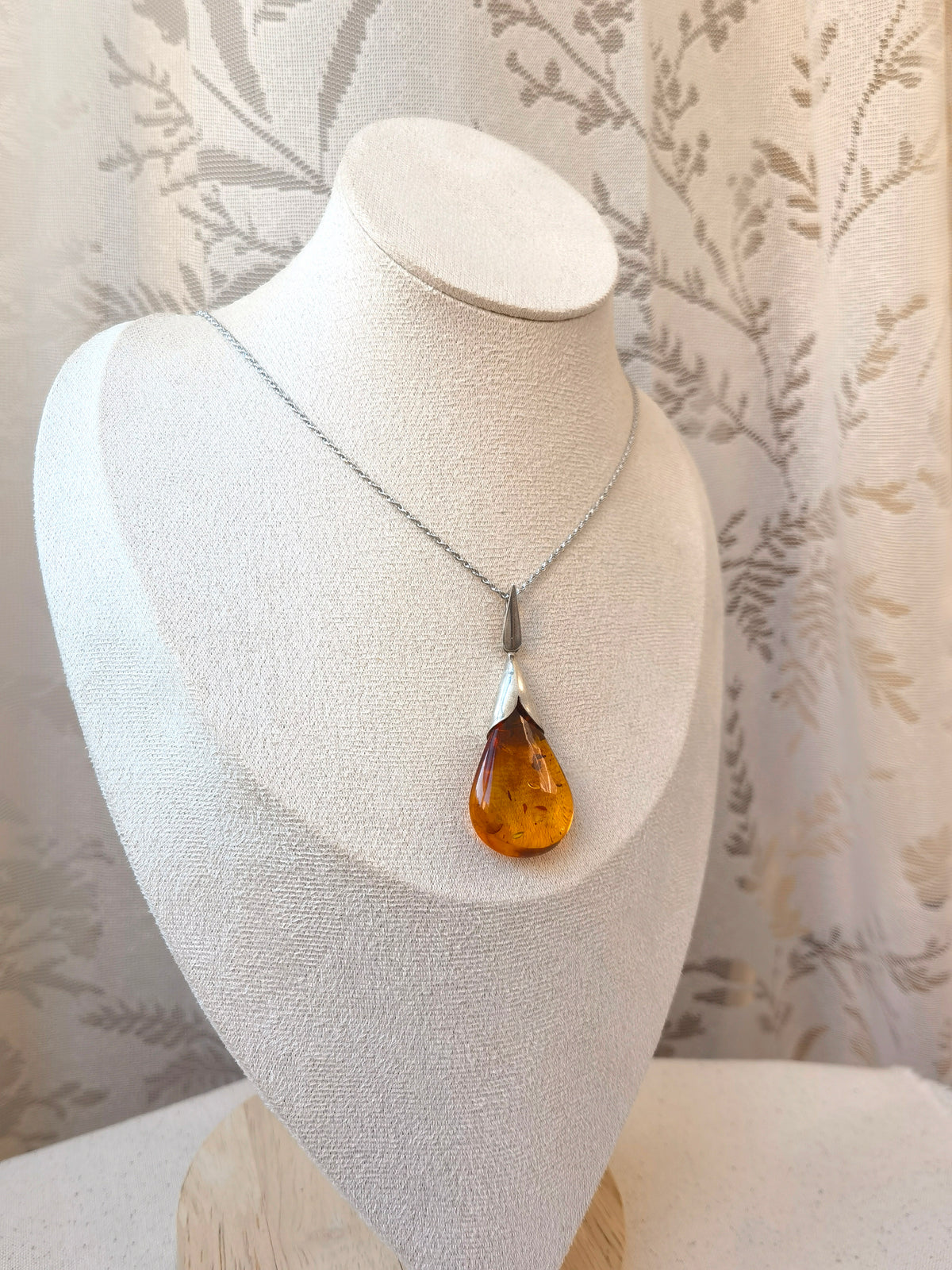 Natural Floral Amber & Matte Silver Necklace — Necklace with a teardrop-shaped amber pendant on a white bust against a